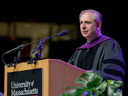 man standing at a podium that says University of Massachusetts Amherst wearing a black robe with a purple stole