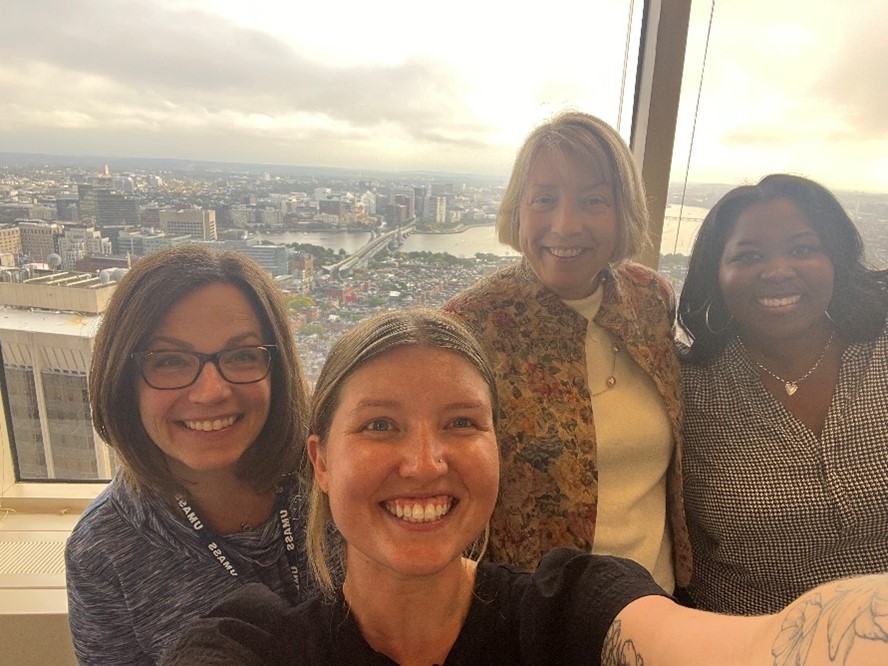 Four women smiling in a selfie with the Boston skyline behind them.