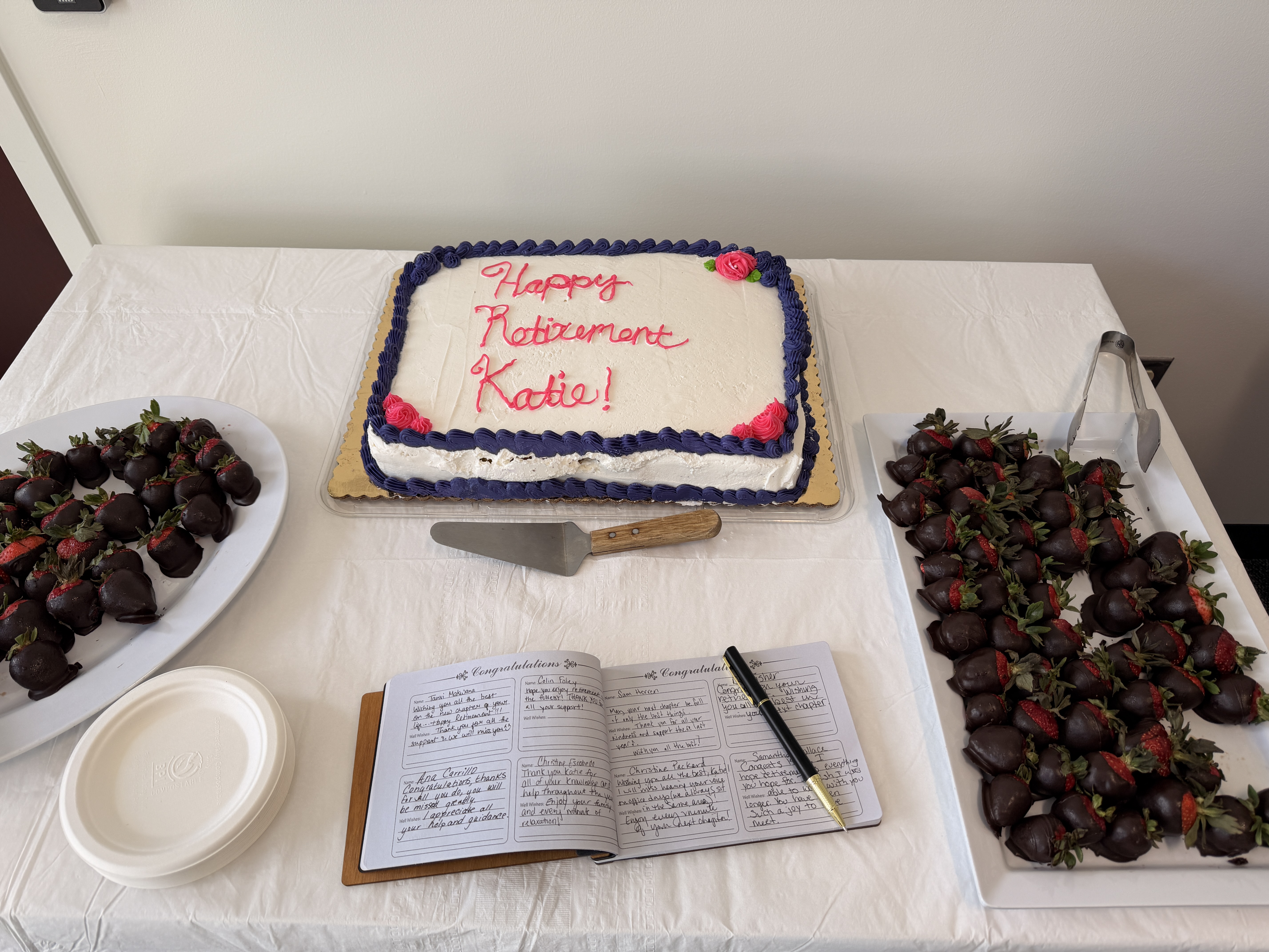 Table with white tablecloth with a cake with Happy Retirement Katie written in icing surrounded by chocolate covered strawberries and paper with handwritten notes. 