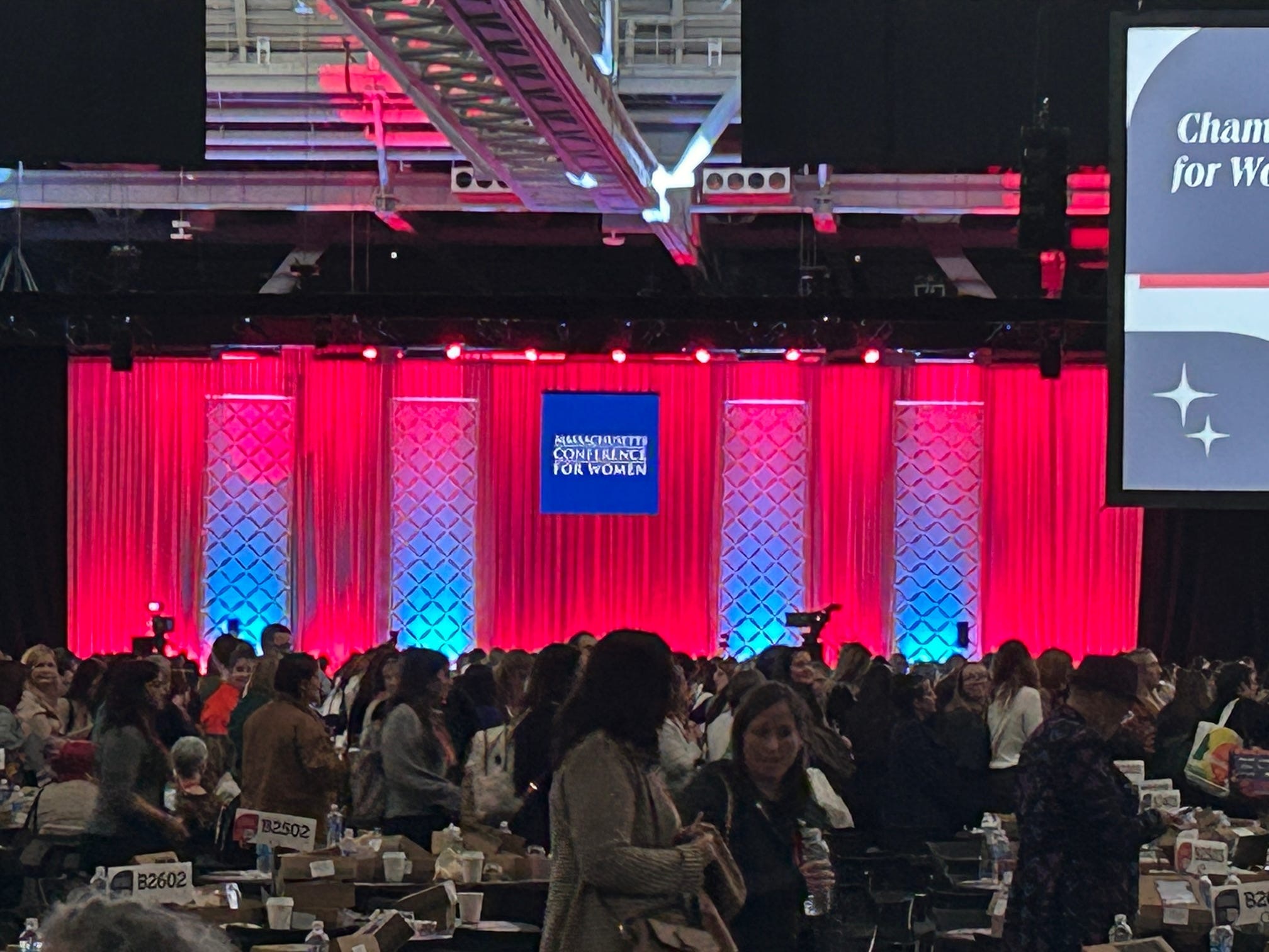 Conference Hall with many people standing around tables, a red background with a blue Massachusetts Conference for Women logo