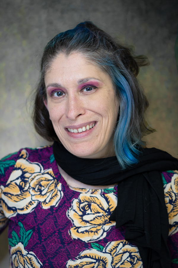 A light-skinned woman with brown shoulder-length hair with blue highlights smiles at the camera. She wears a black scarf and a floral shirt.