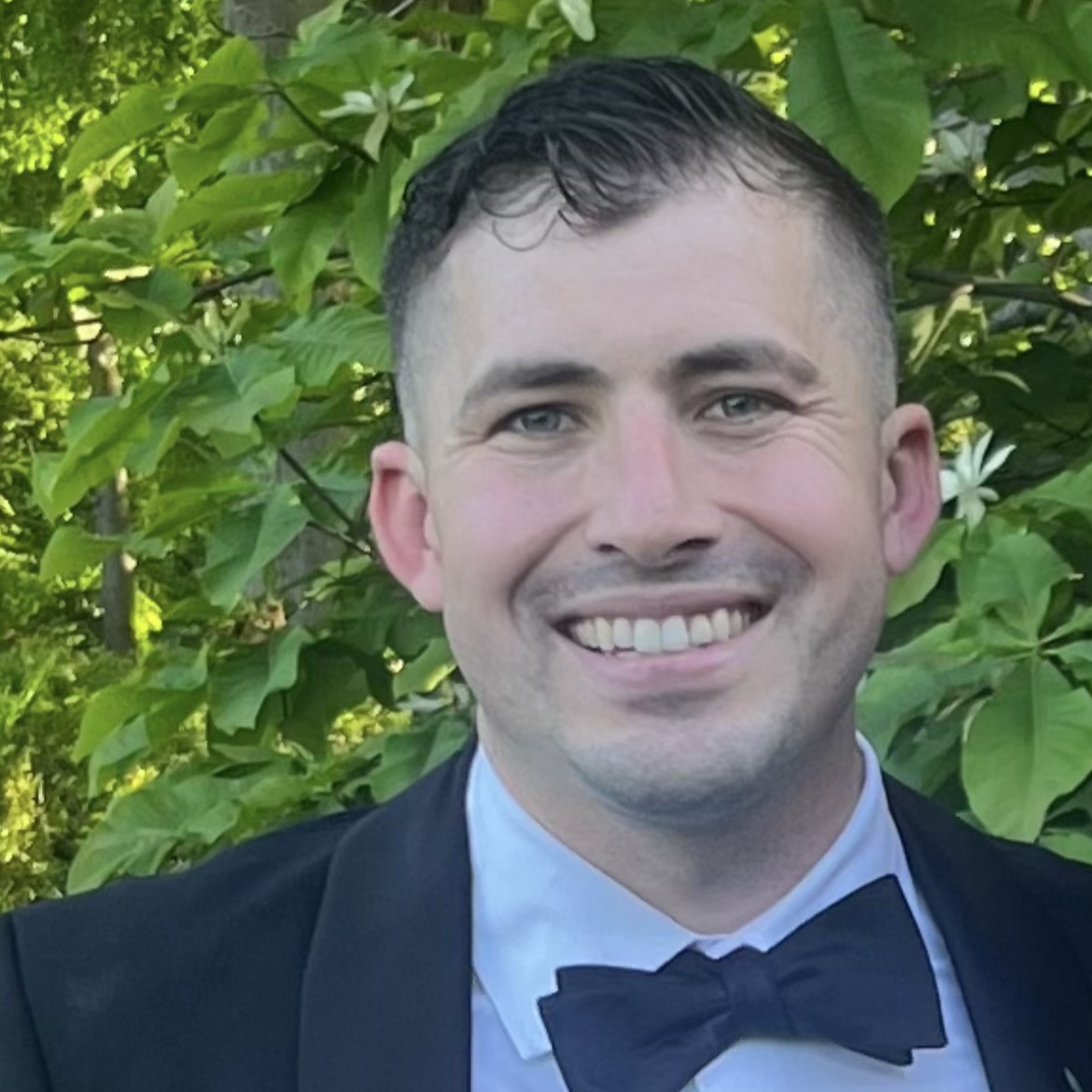 white male with black hair smiles while wearing a black blazer, white dress shirt, and black bowtie