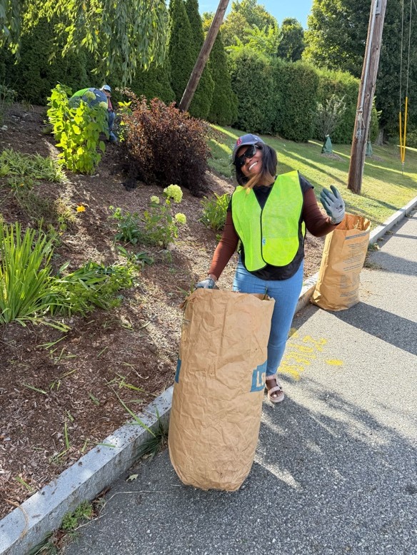 Oriana waves with a garden glove and smiles. She wears a bright green gardening vest and stands behind a gardening bag