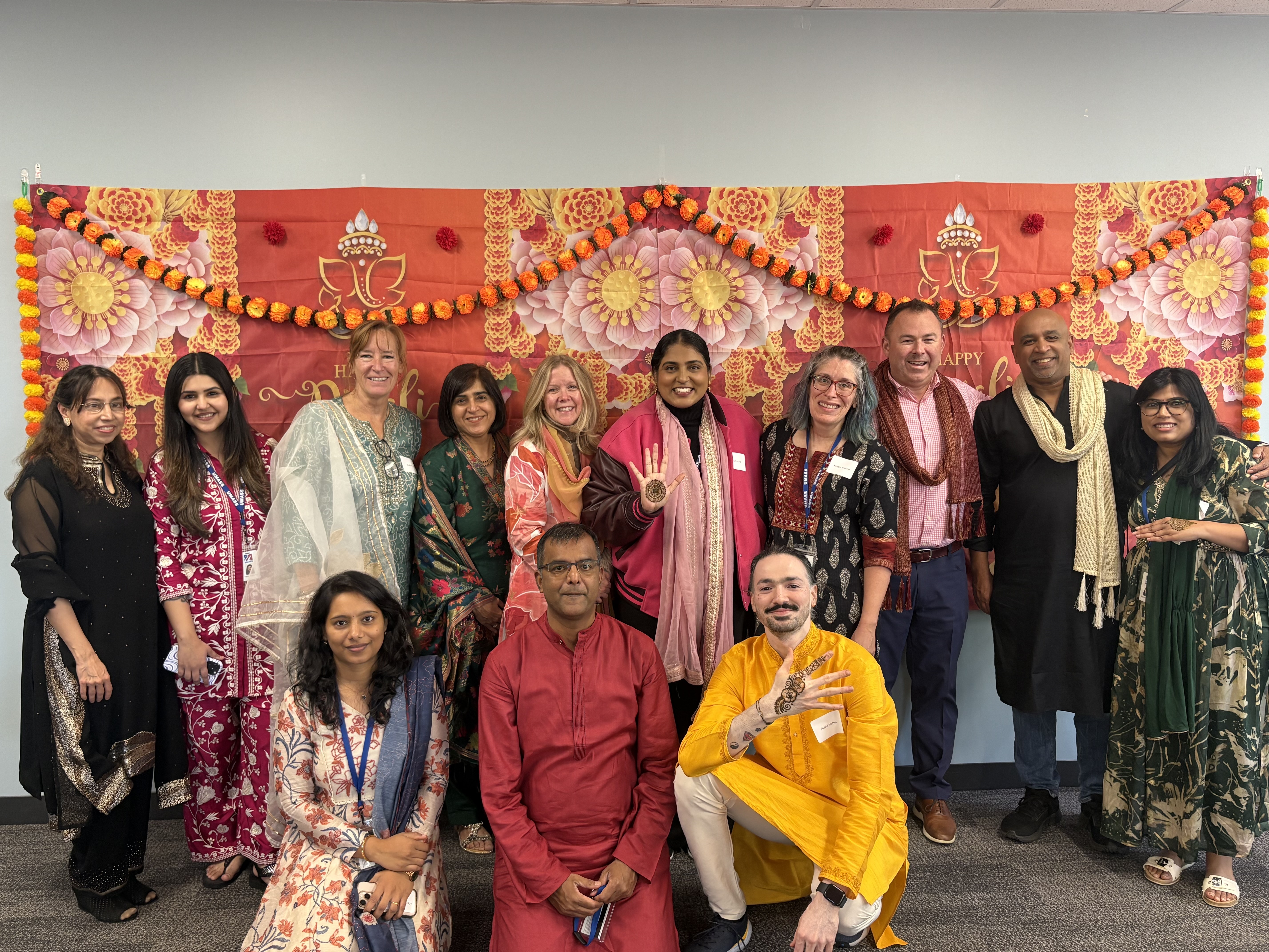 group of people standing together smiling in front of a diwali background all dressed in traditional garments