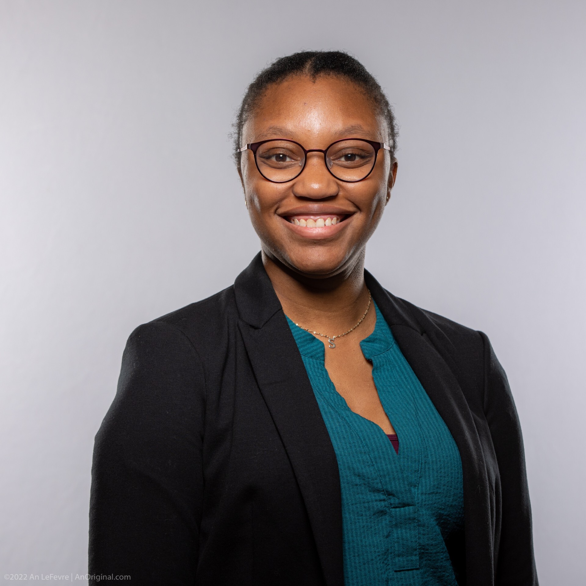 dark-skinned woman with black framed glasses, a teal blouse, and black blazer smiling for her headshot
