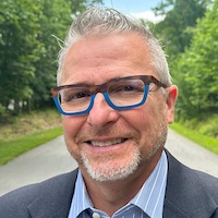 C. Edward Watson, a middle aged man with pale skin and white hair, smiling, wearing glasses, in a grey suit and striped blue shirt