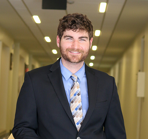 A white, masculine-presenting individual with a brown beard that covers the majority of their face is dressed in a suit and tie and smiles at the camera. They are located in a hallway within the College of Education at the University of Massachusetts Amherst. 