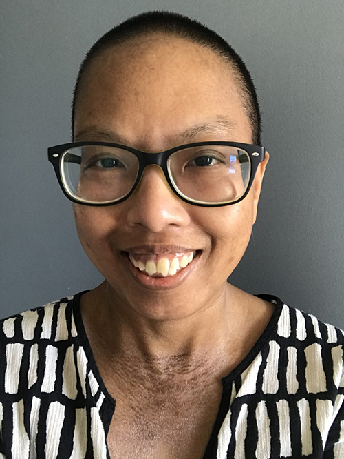 liz indoors with shaved black hair, black eyes, and thick charcoal rimmed glasses. They're wearing a lightweight tunic top that is beige with a black block pattern with a wide v-neck showing their textured darkened skin.