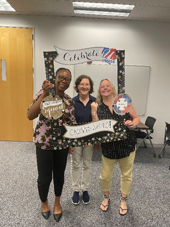 Amanda, a black woman with pulled back hair and glasses, Kathy, a white woman with shoulder length curly hair and glasses, and Meghan Portmann, a white woman with long wavy hair, smile while holding an anniversary cardboard frame.