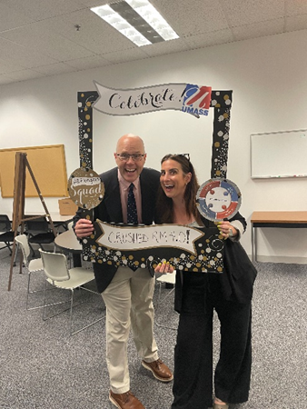 Mike, a white man with a shaved head and glasses and Amy, a white woman with long straight hair, pose with a cardboard anniversary frame. They both make silly wide-mouthed grins.