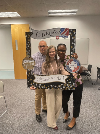 Brian, a white man with shaved hair and glasses, Anna, a white woman with long brown straight hair, and Amanda, a black woman with pulled back dark brown hair and glasses, smile as they hold a cardboard anniversary frame.