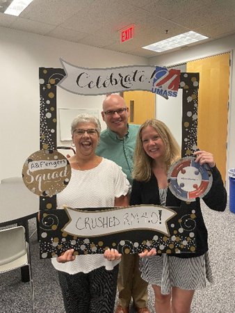Carol, a white woman with short curl gray hair, Jason, a white man with glasses and a shaved head, and Jacquie, a a white woman with medium length blonde hair, smile and pose with a cardboard anniversary frame.