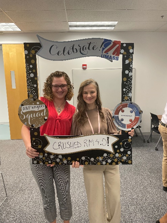 Deirdre, a white woman with short curly hair, and Anna, a white woman with long hair smile as they pose through a cardboard anniversary frame.