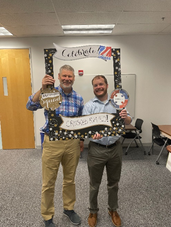 Jeff, a white man with gray hair and a bear, and Patrick, a man with ginger colored hair and a beard, smile while posing with the cardboard anniversary frame.