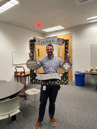 A white man with short brown hair wears pink heart shaped party glasses and poses holding a cardboard anniversary frame.