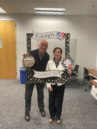 John, a white man with a shaved head, and Lisa, a white woman with pulled back dark brown hair, smile and pose with a cardboard anniversary frame.