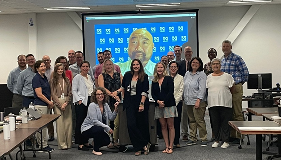 A large group of people stand for a photo with another person zooming in on large screen behind them.