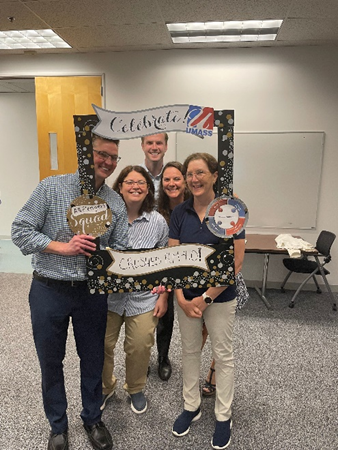 Andrew, a white man with glasses and short brown hair, Monica, a white woman with short curly hair brown and glasses, Sean, a white man with short hair, Kathey, a white woman with long brown wavy hair, and Kathy, a white woman with shoulder length brown curly hair and glasses, pose in a cardboard anniversary frame they are holding up.