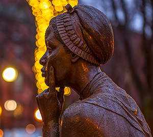 A statue of a woman deep in thought with her finger pressed against her face. The statue is covered in rain and is illuminated by holiday lights in the background. 