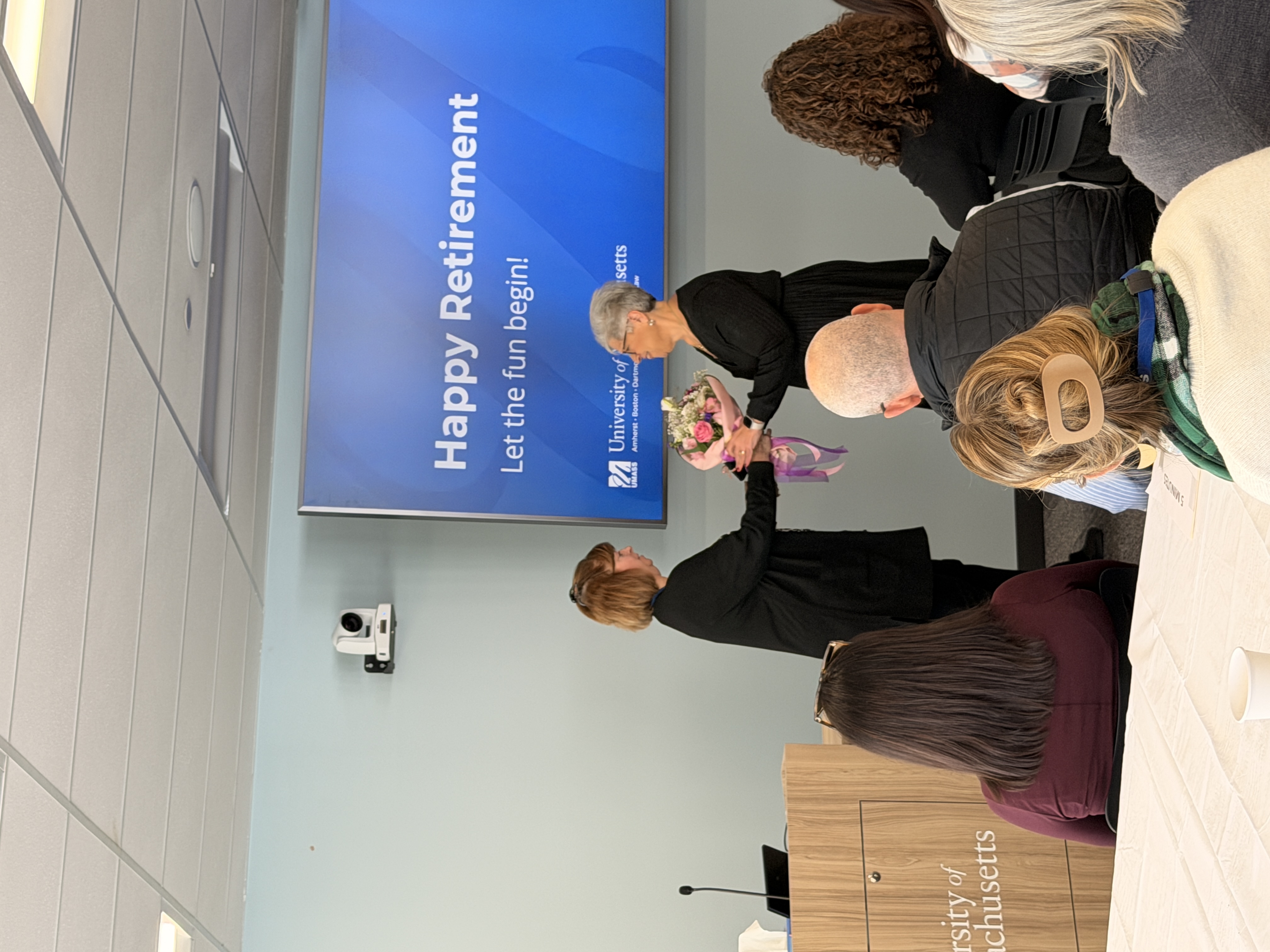 Carol Dugard standing in front of a blue monitor that says Happy Retirement, receiving flowers from Diane Pelletier