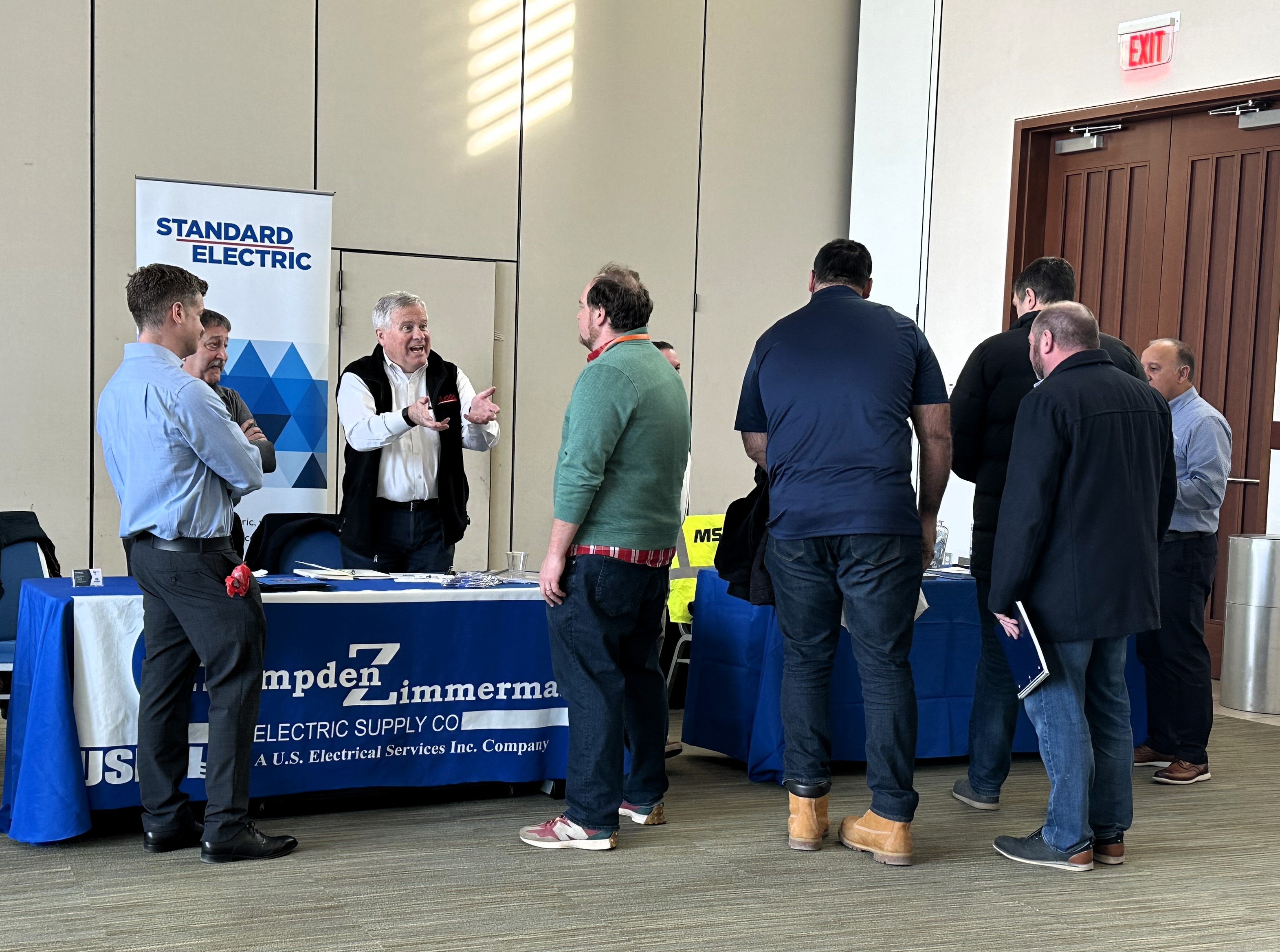 Men gathered around a waist height table with a blue tablecloth listening to a man talking to them. Behind him is a vertical banner saying "Standard Electric"