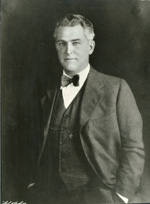 A light-skinned man with gray and white hair and a shaven face stands with one hand in a pant pocket, staring at the camera. He wears a dark suite coat, white shirt, and a bow tie.