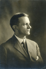 A middle-aged light-skinned man with dark hair combed back and a shaven face sits looking away from the camera. He wears a light gray suit coat, white shirt, a tie, and has a handkerchief in his suit coat pocket.