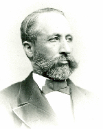 A light-skinned man with dark receding hair and a dark beard poses firmly in a bow tie, white shirt, and suit coat.