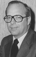 A light-skinned man with gray hair and a cleanly shaven face wears glasses. He smiles into the camera, wearing a gray suit coat, a dark tie, and a white shirt.