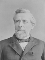 A light-skinned man with white hair and a white beard stares softly off camera. He wears a dark gray coat and a white shirt.