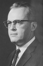 A light-skinned man with white-gray hair wears glasses and is cleanly shaven. He stares off camera, wearing a black suit coat, a dark tie, and a white shirt.