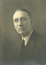 A middle-aged light-skinned man with combed back hair and a shaven face, wearing glasses, stares directly into the camera. He wears a dark suit coat, dark tie, and a white shirt.