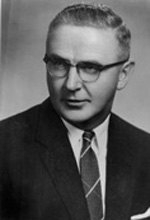 A light-skinned middle aged man with medium toned hair wears glasses and is cleanly shaven. He looks off camera and wears a black suit coat, a dark tie, and a white shirt.