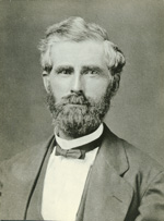 A light-skinned man with white-gray hair and a dark beard stares softly into the camera. He wears a bow tie, white shirt, and suit coat.