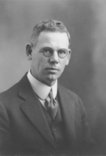 An older light-skinned man with gray hair wears glasses and stares directly into the camera. He wears a suit coat, tie, and a white shirt.