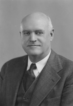 A light-skinned man with balding white hair and a cleanly shaved face smiles into the camera. He wears a dark suit coat, a dark tie, and a white shirt.