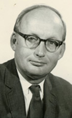 A light-skinned man with balding gray hair and a cleanly shaven face wears glasses. He stares off camera, wearing a dark gray suit coat, a dark tie, and white shirt.