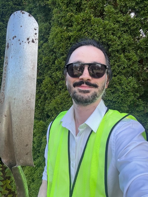 Akshit wears sunglasses and a bright green gardening vest. He smiles while taking a selfie holding a metal shovel.