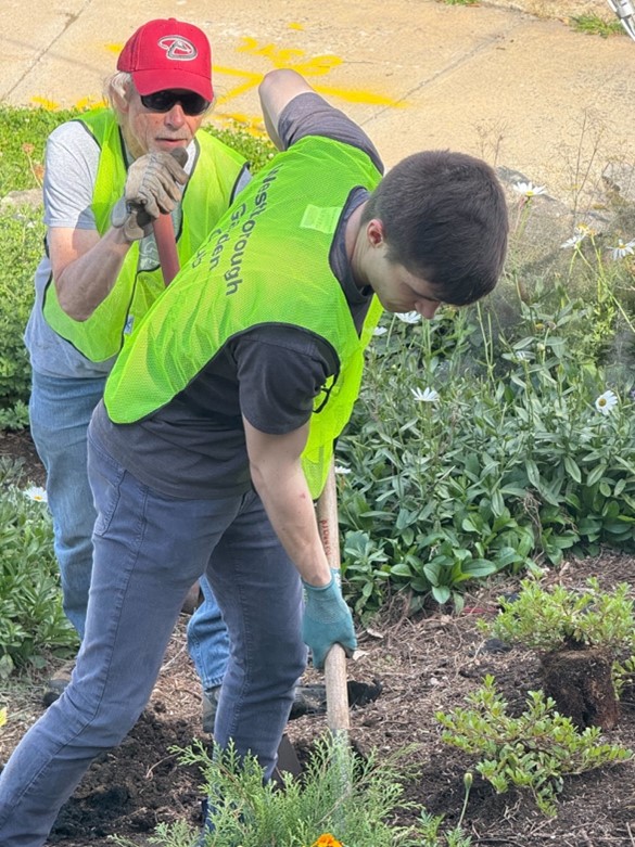 Jake pushes a shovel into the ground as he leans forward wearing a bright green gardening vest. Another person stands behind him watching him dig.