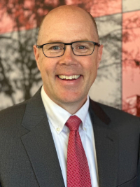 a light skinned balding man with glasses wearing a dark suit and a red tie, smiling