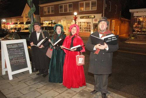 Four carolers sing on a side walk, dressed in old fashioned dresses and suits.