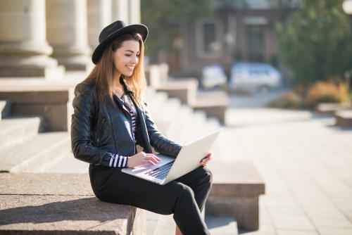A young light-skinned woman sits on the marble steps of a building workin on her laptop. Some trees and brick buildings can be seen in the distance.