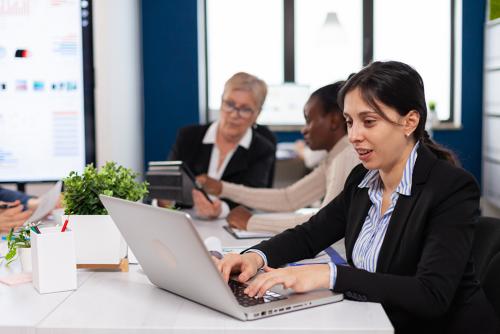 Two light-skinned women and one dark-skinned woman sit at a conference table working.