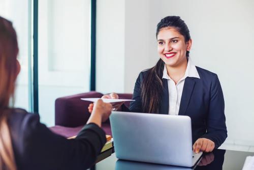 A smiling medium-light skin-toned woman in business attire sits in front of a computer and accepts a letter from another medium skin-toned woman.