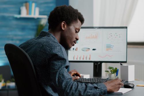 A dark-skinned man sits at a desk taking notes in a notebook. His computer monitor displays a dashboard.