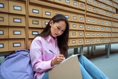 college student in a pink shirt and jeans sitting on the floor in a library writing in a notebook