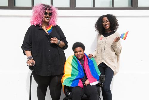 Three Black and disabled folx decked out happily in rainbow pride flags. On the left, a non-binary person holds a cane in one hand and waves a mini flag in the other. In the middle, a non-binary person sits in their power wheelchair, draped in a large flag. On the right, a femme rests one hand on their friend's shoulder and waves a mini flag around with the other hand.