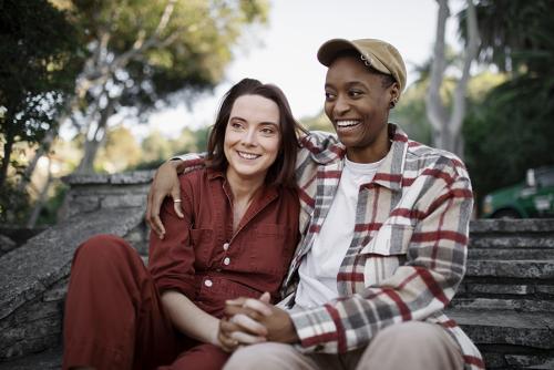 A light-skinned woman and dark-skinned masculine-presenting woman sit together with their hands clasped.