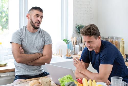 A medium-skinned man and light-skinned man talk in their kitchen.