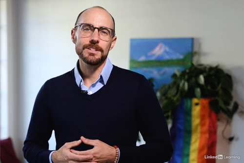 A light skinned man presents with the Pride flag behind him.